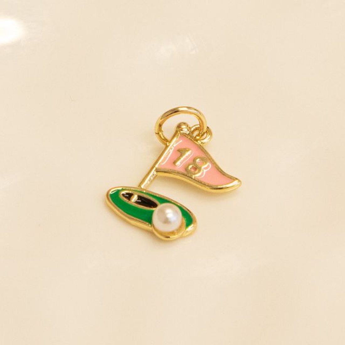 A gold golf charm on a white dish: a pink 18th-hole flag on a green with a pearl golf ball.