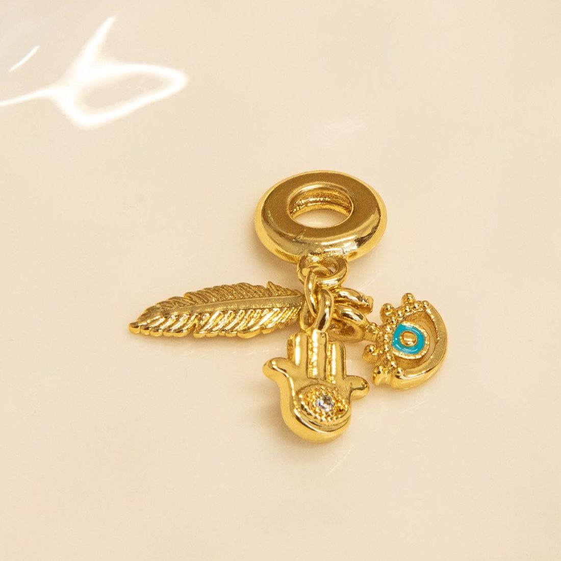 Gold charms on a white dish: a feather, a hamsa hand and an evil eye with turquoise enamel.