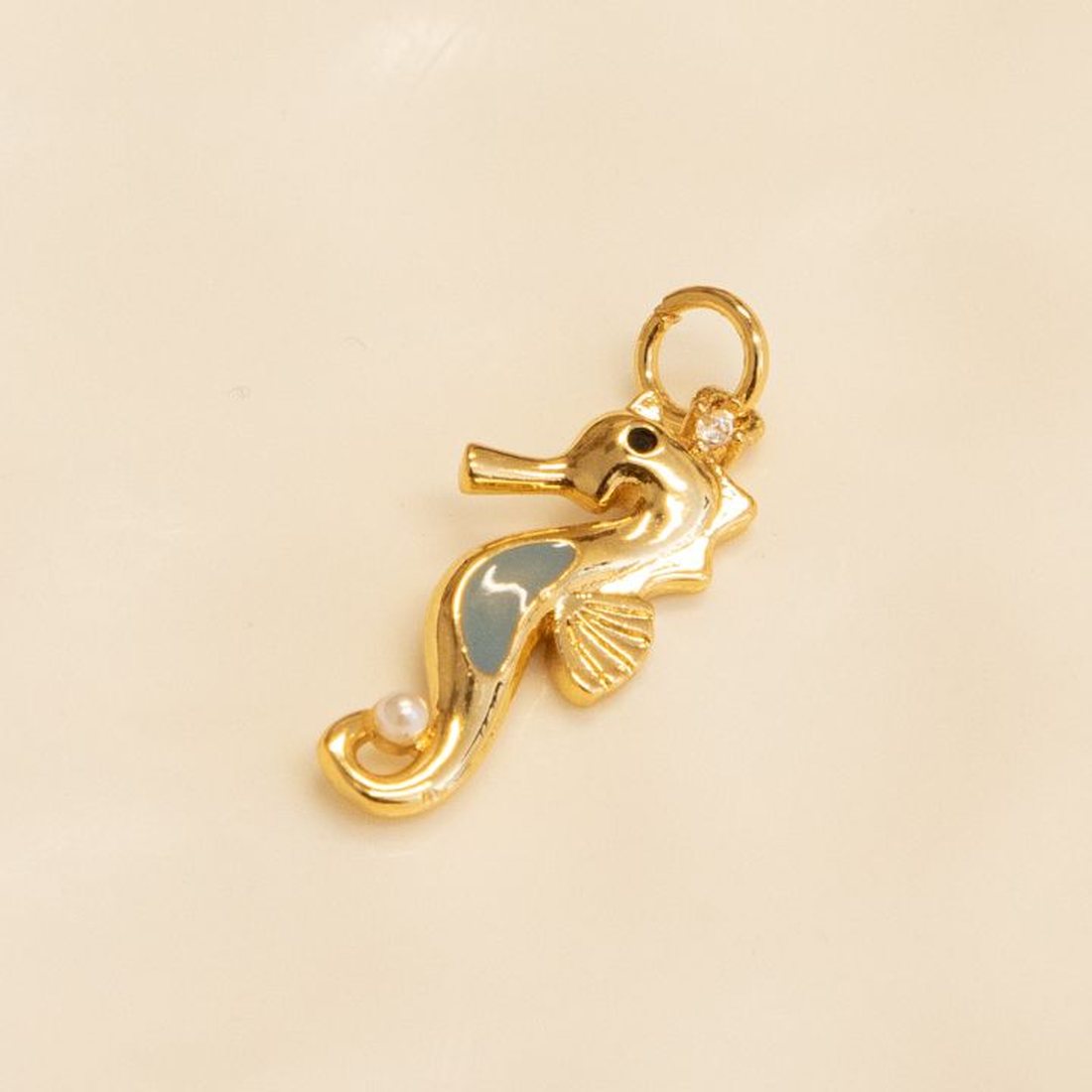 A gold seahorse charm with soft blue enamel and a tiny pearl, on a white dish.