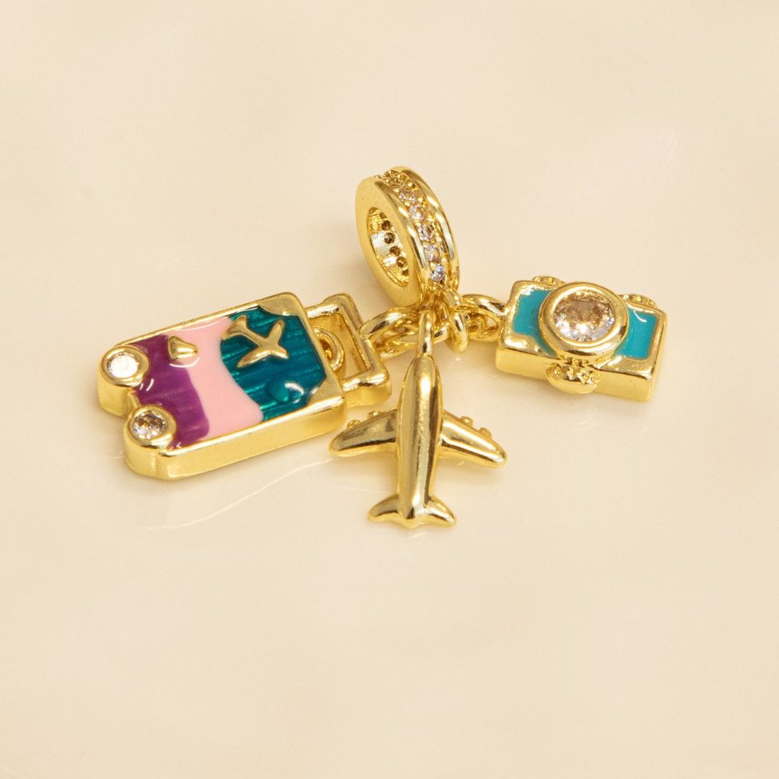 Gold travel charms on a white dish: a suitcase in pink, purple and teal enamel, an airplane and a turquoise camera.
