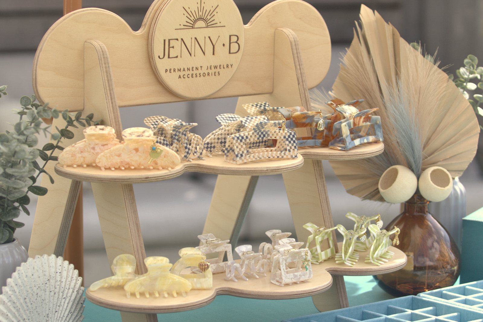 A wooden Jenny B display stand with two shelves of claw clips in checkered, marbled and soft pastel colors, beside dried palm leaves.
