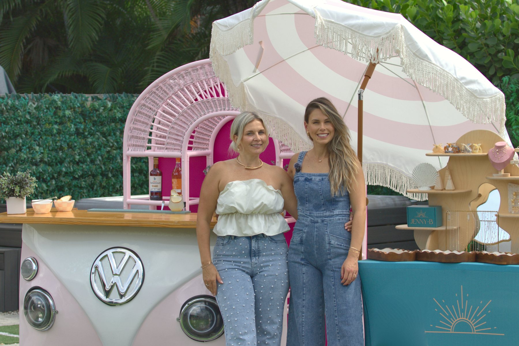 Jenny and Joanna side by side between the pink VW Dream Bar and the Jenny B Charm Bar, under a pink and white fringe umbrella.