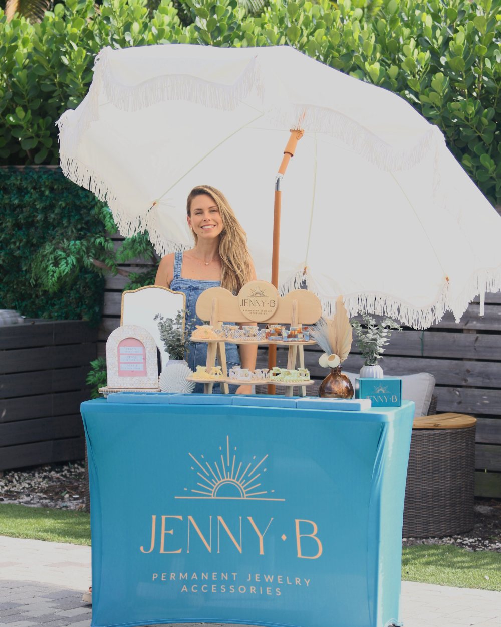 A smiling host behind the teal Jenny B pop-up table, set with chains and charms under a white fringe umbrella.