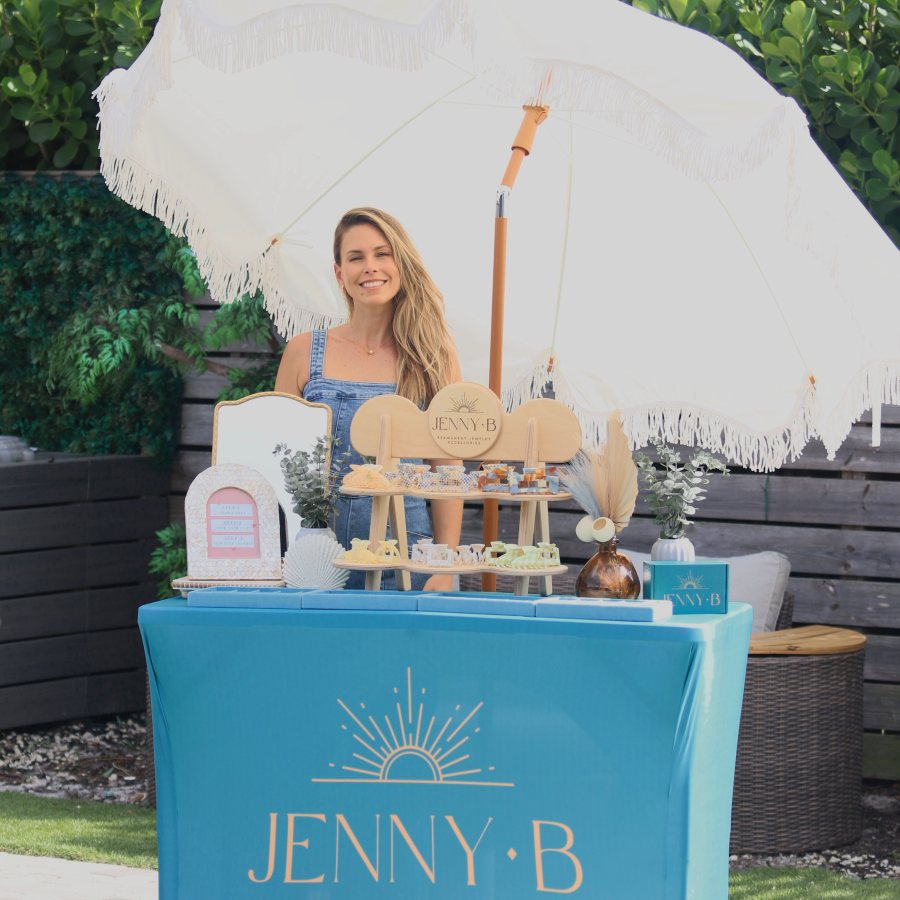 A smiling host behind the teal Jenny B pop-up table, under a white fringe umbrella.