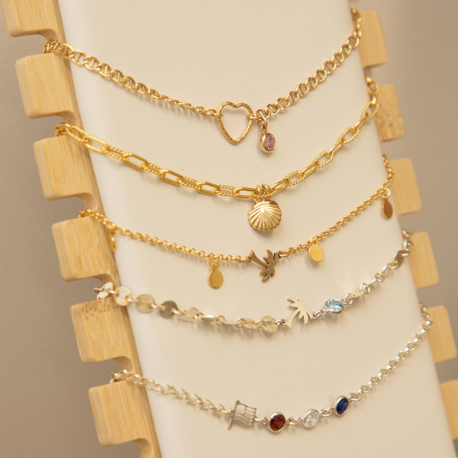 Five bracelets on a display stand: gold chains with a heart, a shell and a palm tree, and silver chains with a palm tree, flag and colored stones.