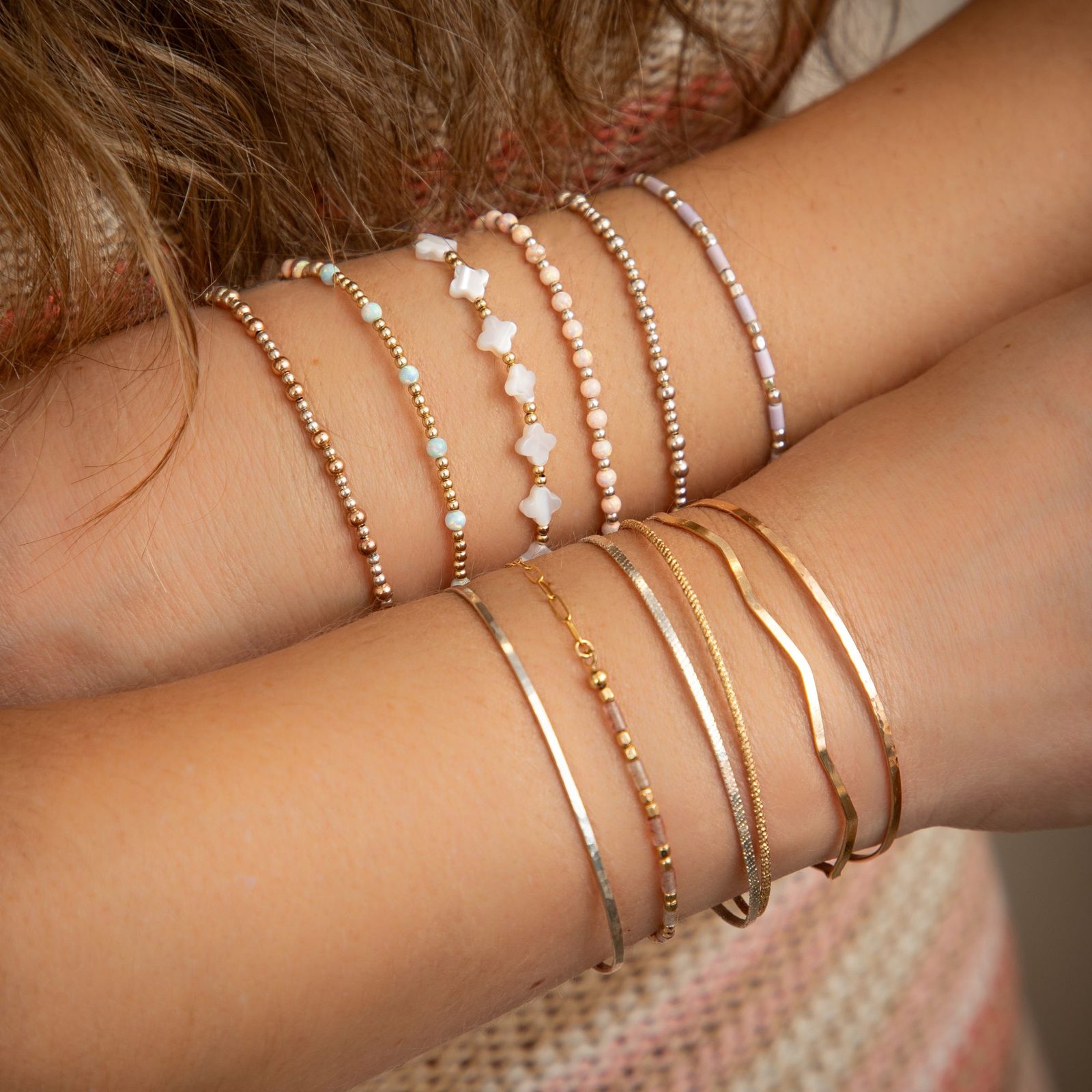 Crossed arms wearing a stack of beaded bracelets with opal, pearl and mother-of-pearl clover beads, above gold-filled bangles and chains.