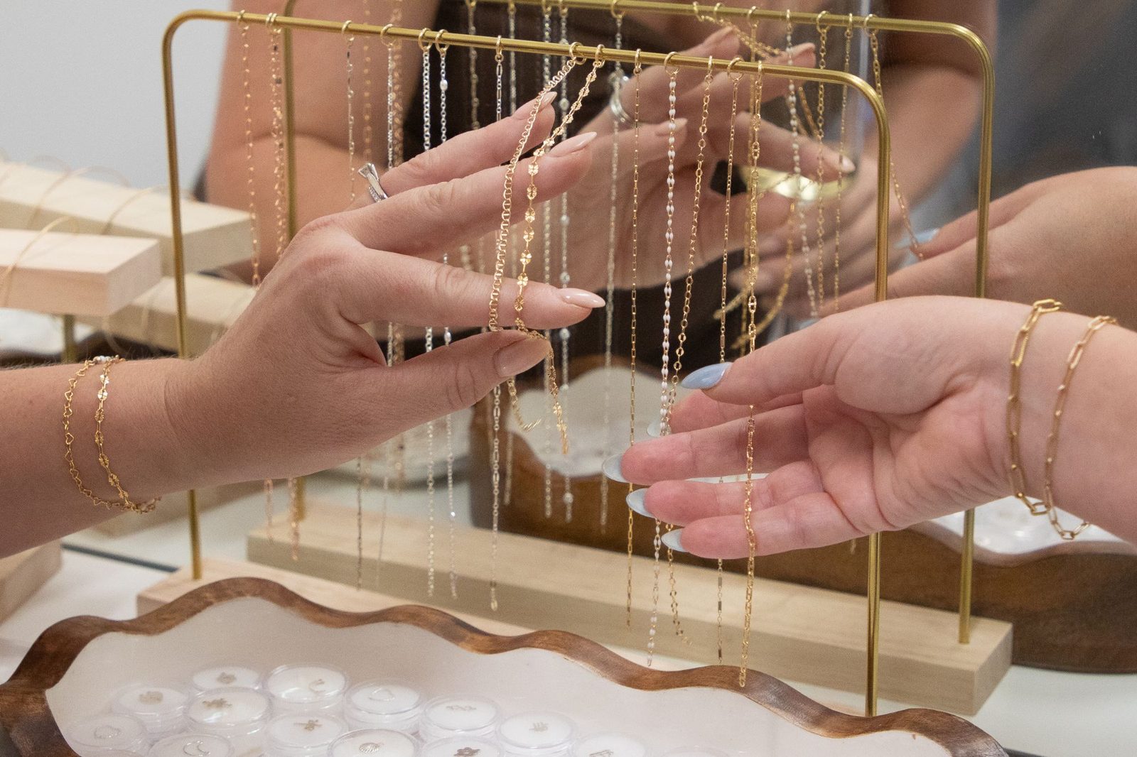 Two hands choosing gold chains from a brass rack, above a wavy wooden tray of charms.