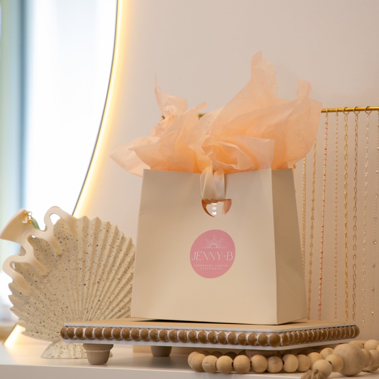 A cream Jenny B gift bag with peach tissue paper, a shell-shaped claw clip and a rack of gold chains.