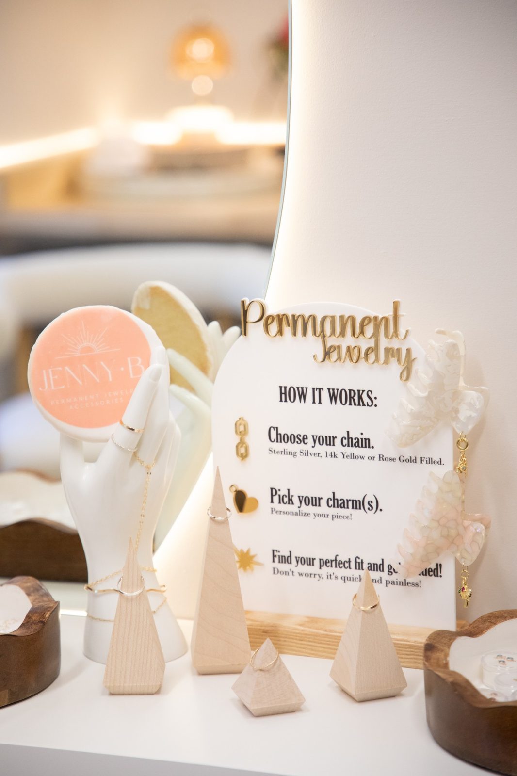 A &ldquo;Permanent Jewelry, How it works&rdquo; sign beside a ceramic hand wearing gold rings and a hand chain, wooden ring cones and claw clips with charms.
