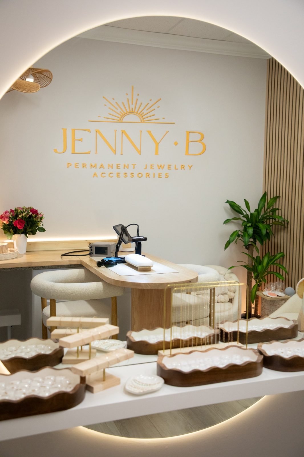 The round backlit mirror reflecting the gold Jenny B logo, the welding table and trays of charms.